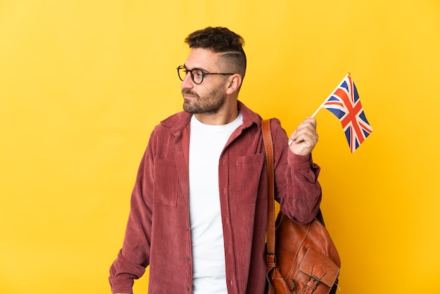 Kaukasischer Mann, der eine Flagge des Vereinigten Königreichs lokalisiert auf gelber Wand hält, die zur Seite schaut