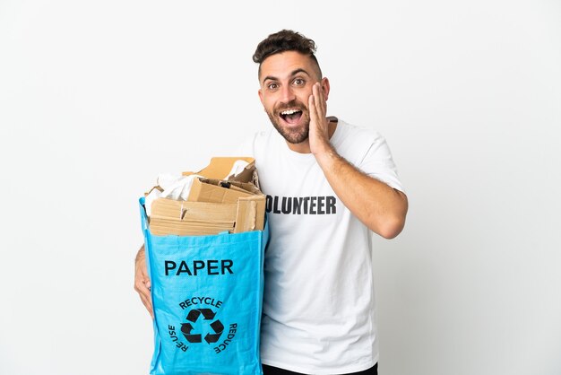 Kaukasischer Mann, der eine Recycling-Tasche voller Papier hält, um sie isoliert auf weißem Hintergrund mit Überraschung und schockiertem Gesichtsausdruck zu recyceln