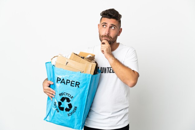 Kaukasischer Mann, der eine Recyclingtüte voller Papier hält, um isoliert auf weißem Hintergrund zu recyceln, der Zweifel hat und denkt