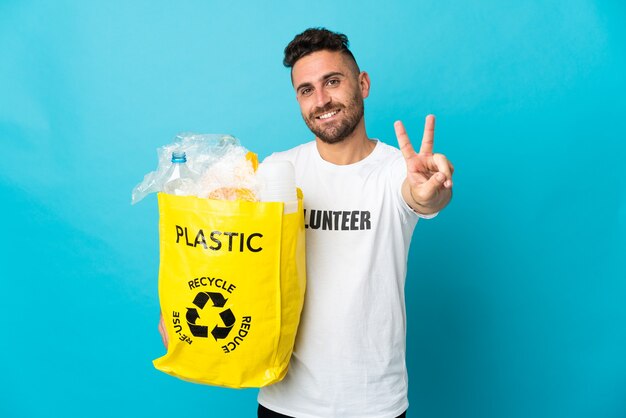 Kaukasischer Mann, der eine Tasche voller Plastikflaschen hält, um auf blau lächelnd und siegreiches Zeichen isoliert zu recyceln