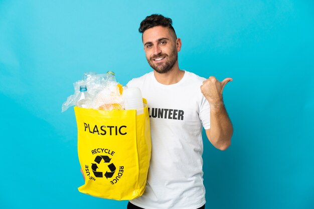 Kaukasischer Mann, der eine Tasche voller Plastikflaschen hält, um isoliert auf der blauen Wand zu recyceln, die zur Seite zeigt, um ein Produkt zu präsentieren