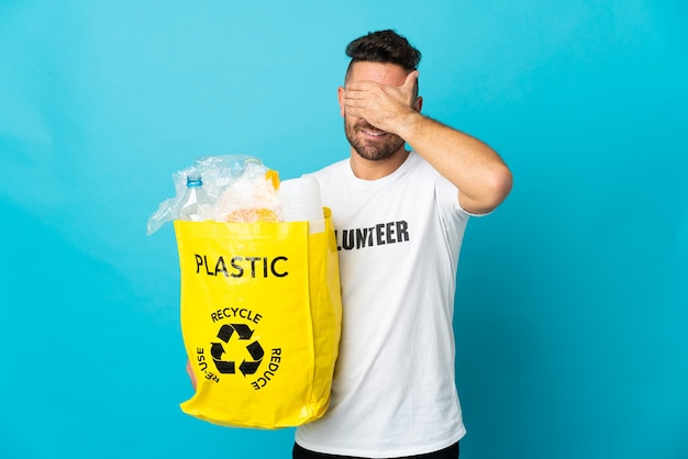 Kaukasischer Mann, der eine Tasche voller Plastikflaschen zum Recyceln hält, isoliert auf blauem Hintergrund, der die Augen mit den Händen bedeckt. Will etwas nicht sehen