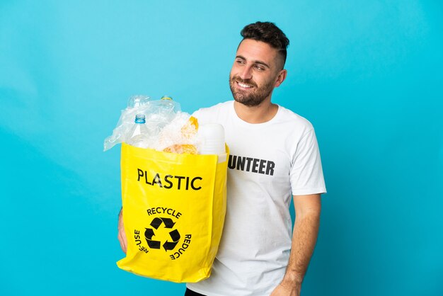 Kaukasischer Mann, der eine Tasche voller Plastikflaschen zum Recyceln hält, isoliert auf blauem Hintergrund, der zur Seite schaut und lächelt