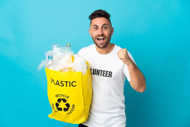 Kaukasischer Mann, der eine Tasche voller Plastikflaschen zum Recyceln hält, isoliert auf blauem Hintergrund, feiert einen Sieg in Siegerposition