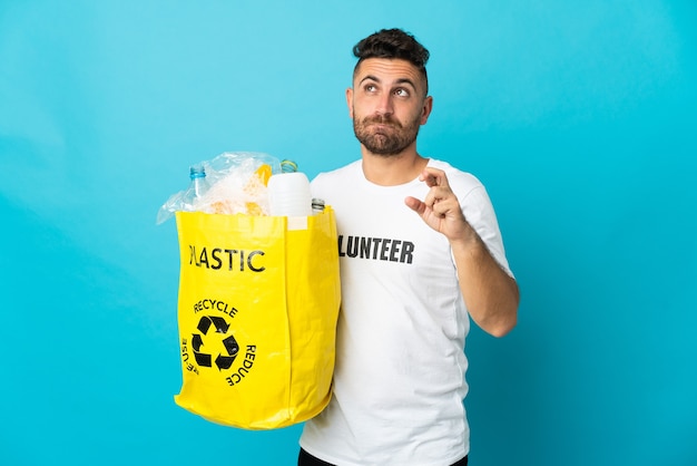 Kaukasischer Mann, der eine Tasche voller Plastikflaschen zum Recyceln hält, isoliert auf blauem Hintergrund, mit gekreuzten Fingern und das Beste wünscht