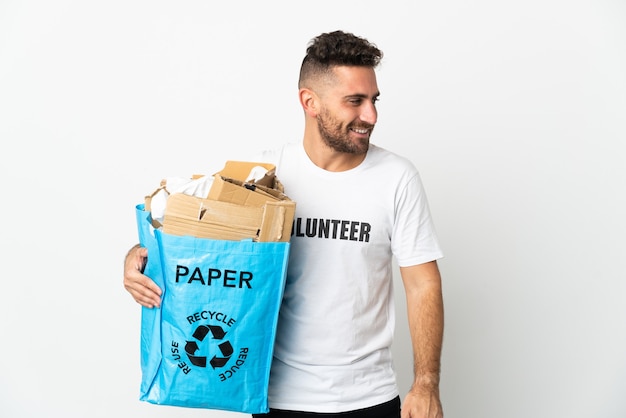 Kaukasischer Mann, der einen Recyclingbeutel voll Papier hält, um lokalisiert auf weißem Blick zur Seite und lächelnd zu recyceln