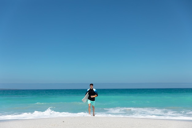 Kaukasischer Mann, der mit einem Surfbrett aus dem Meer an den Strand geht, mit blauem Himmel und Meer im Hintergrund. Surfen in einem gesunden, aktiven Strandurlaub