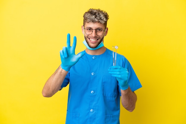 Kaukasischer Mann des Zahnarztes, der Werkzeuge lokalisiert auf gelbem Hintergrund glücklich hält und drei mit den Fingern zählt