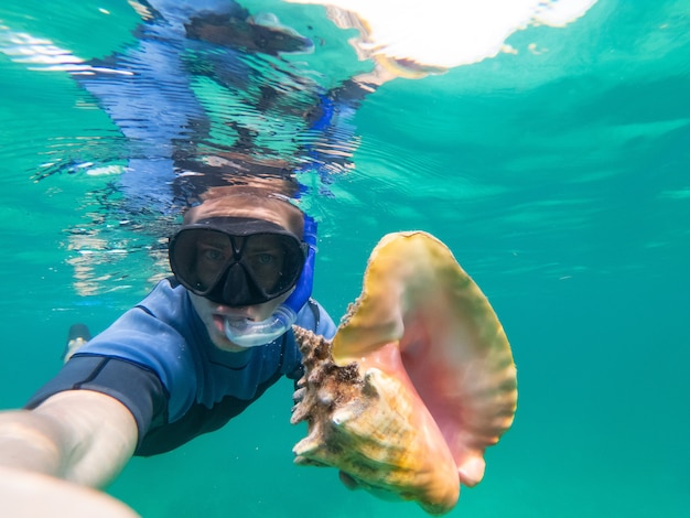 Kaukasischer Mann hält und riesige Muschel beim Schnorcheln unter Wasser. Reise-, Urlaubs- und Abenteuerkonzept.