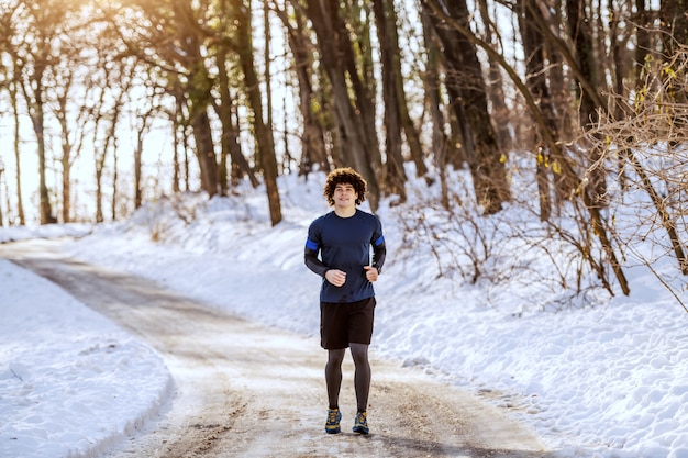 Kaukasischer Mann in voller Länge in Sportbekleidung und mit lockigem Haar auf der Landstraße. Winter. Outdoor-Fitness-Konzept.