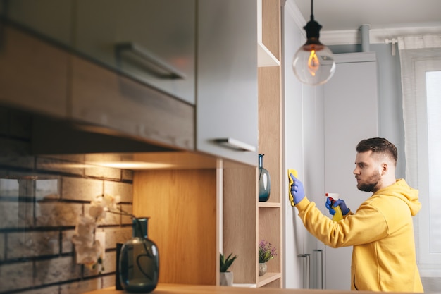 Kaukasischer Mann mit Bart, der einen gelben Pullover trägt, putzt das Haus mit Handschuhen und Spray using