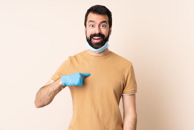 Kaukasischer Mann mit Bart, der mit einer Maske und Handschuhen über isolierter Wand mit überraschendem Gesichtsausdruck schützt