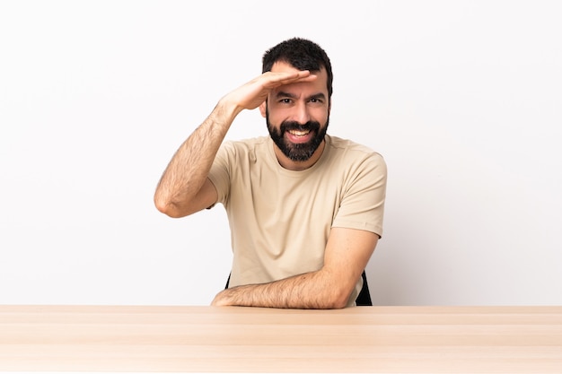 Kaukasischer Mann mit Bart in einem Tisch, der weit weg mit der Hand schaut, um etwas zu schauen