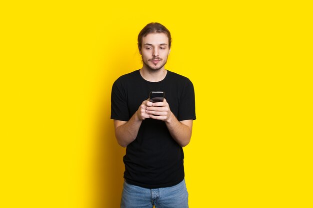 Kaukasischer Mann mit Bart und langen Haaren plaudert auf Handy auf einer gelben Studiowand