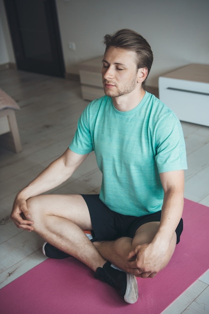 Kaukasischer Mann mit blonden Haaren meditiert auf dem Boden, trägt Sportbekleidung und benutzt Yoga-Teppich