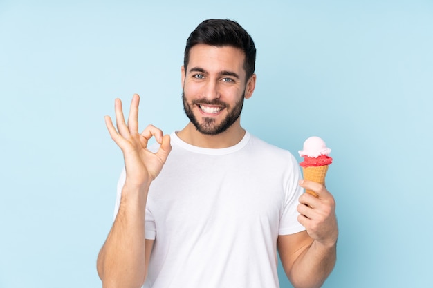 Kaukasischer Mann mit einem Kornetteis lokalisiert auf blauer Wand, die ok Zeichen mit Fingern zeigt