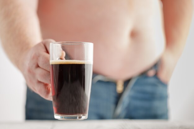 Kaukasischer Mann mit fettem Bierbauch, der ein Glas dunkles Ale hält. Jeans sind sehr eng