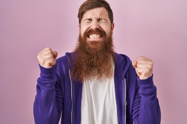 Kaukasischer Mann mit langem Bart, der über rosafarbenem Hintergrund steht und mit erhobenen Armen und geschlossenen Augen auf den Erfolg freut und den Sieg lächelnd feiert. Gewinner-Konzept.