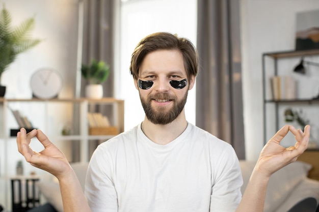 Foto kaukasischer mann mit schwarzen anti-aging-augenklappen sitzt mit mudra-händen an seinem arbeitsplatz zu hause