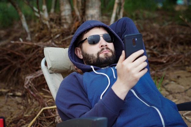 Kaukasischer Mann mit Sonnenbrille ruht sich mit einem Smartphone auf einem Campingstuhl aus.