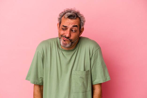 Kaukasischer Mann mittleren Alters, der auf rosafarbenem Hintergrund isoliert ist, zuckt mit den Schultern und öffnet die Augen verwirrt.