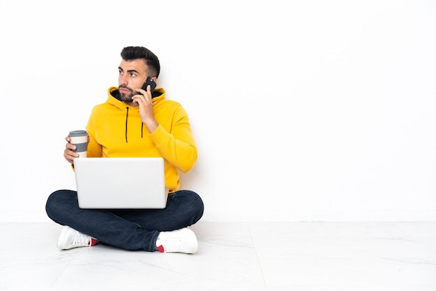 Kaukasischer Mann sitzt auf dem Boden mit seinem Laptop, der Kaffee zum Mitnehmen und ein Handy hält