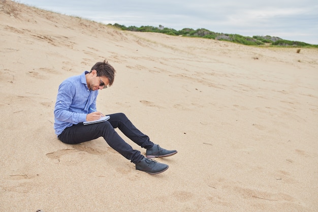 Kaukasischer Mann sitzt auf dem Sand des Strandes an der Küste und schreibt in sein Notizbuch. In formeller Kleidung gekleidet.