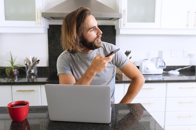 Kaukasischer Mann sitzt mit Laptop in der Küche und spricht mit dem Smartphone