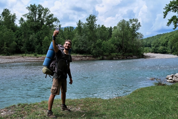 Kaukasischer Mann steht mit großem Wanderrucksack am Ufer des Gebirgsflusses und macht Selfie