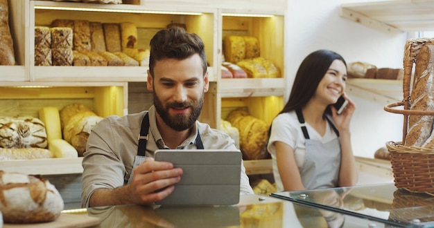 Kaukasischer Mannverkäufer, der Tablet-Gerät verwendet, während er an der Theke in der Bäckerei steht, seine Kollegin, die hinter ihm am Telefon spricht. Innerhalb