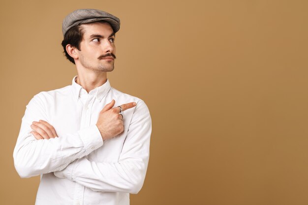 Kaukasischer schnurrbärtiger Mann mit Hut, der mit dem Finger auf Kopienraum zeigt, isoliert auf Beige