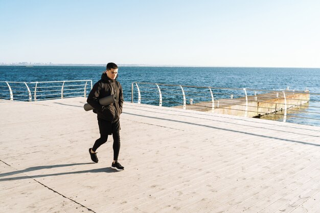 Kaukasischer Sportler 20er Jahre in schwarzer Kleidung mit Ohrstöpseln, die nach dem morgendlichen Training am Meer spazieren gehen