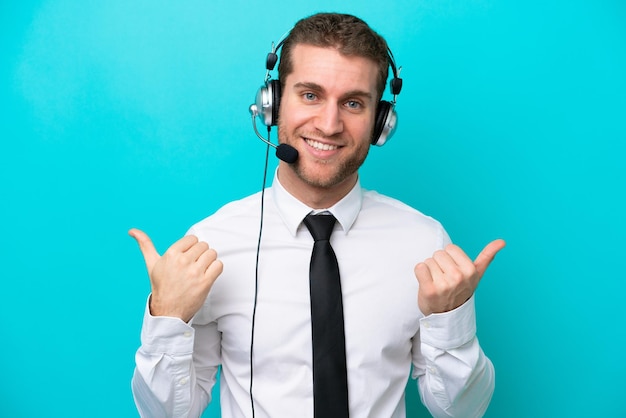 Kaukasischer Telemarketer, der mit einem Headset arbeitet, das auf blauem Hintergrund isoliert ist, mit Daumen nach oben, Geste und Lächeln