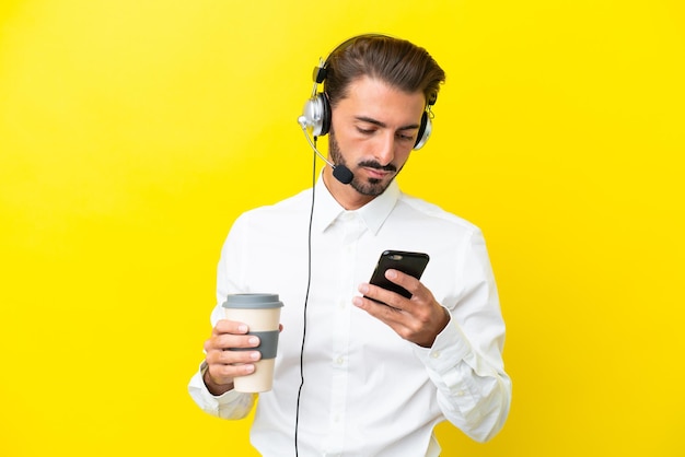 Kaukasischer Telemarketer, der mit einem Headset arbeitet, das auf gelbem Hintergrund isoliert ist und Kaffee zum Mitnehmen und ein Handy hält