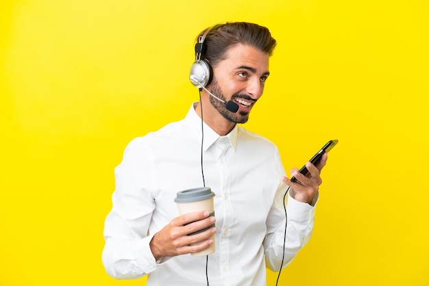 Kaukasischer Telemarketer, der mit einem Headset arbeitet, das auf gelbem Hintergrund isoliert ist und Kaffee zum Mitnehmen und ein Handy hält