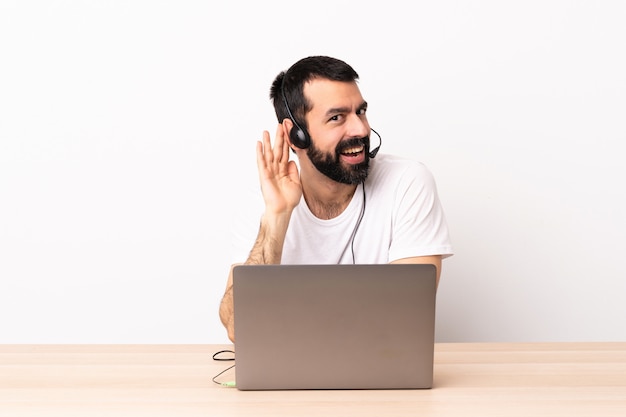 Kaukasischer Telemarketer, der mit einem Headset und mit Laptop arbeitet, der etwas hört, indem man Hand auf das Ohr legt