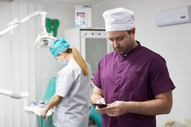 Kaukasischer überzeugter junger männlicher Doktorzahnarzt, der Handy in der zahnmedizinischen Praxis verwendet