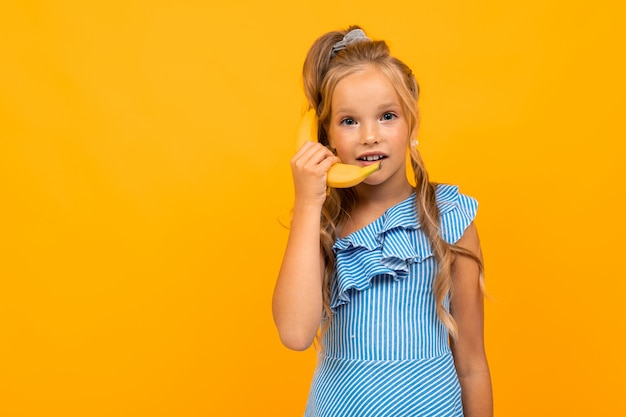 Kaukasisches blondes Mädchen, das eine Banane an einer gelben Wand hält