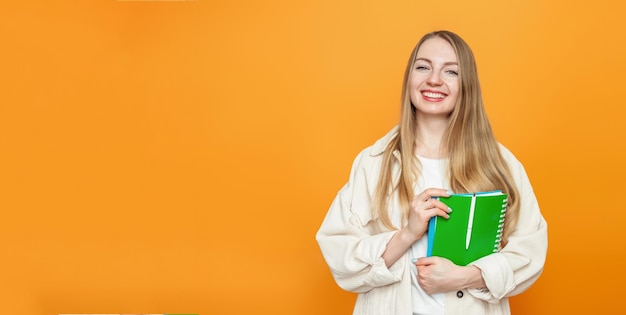 Kaukasisches blondes Studentenmädchen, das ein Buchheft hält und lächelnd in die Kamera schaut, isoliert über orangefarbenem Hintergrund im Studio Kopierraum