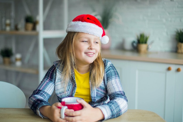 Kaukasisches entzückendes lächelndes Mädchen mit Tee im Weihnachtsmann-Hut in der gemütlichen Küche