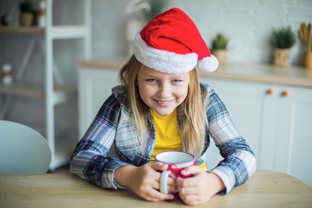 Kaukasisches entzückendes lächelndes Mädchen mit Tee im Weihnachtsmann-Hut in der gemütlichen Küche