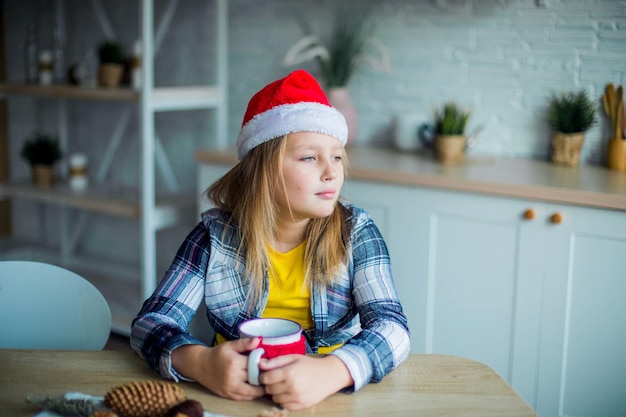 Kaukasisches entzückendes lächelndes Mädchen mit Tee im Weihnachtsmann-Hut in der gemütlichen Küche