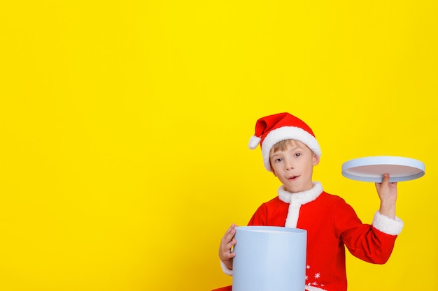 Kaukasisches glückliches süßes Kind, das als Weihnachtsmann verkleidet ist, hält eine offene runde blaue Geschenkbox in einer Hand