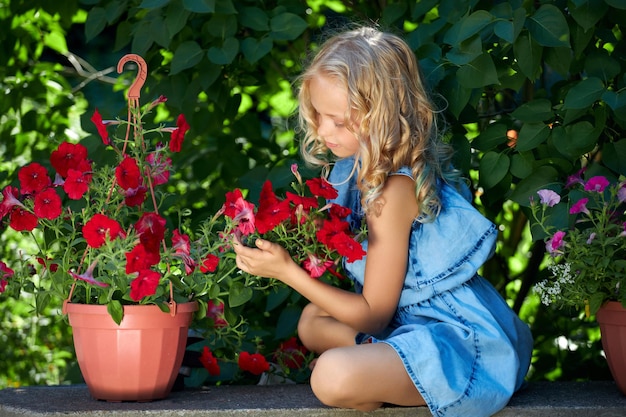 Kaukasisches junges blondes Mädchen in einem blauen Kleid kümmert sich um Blumen im Garten