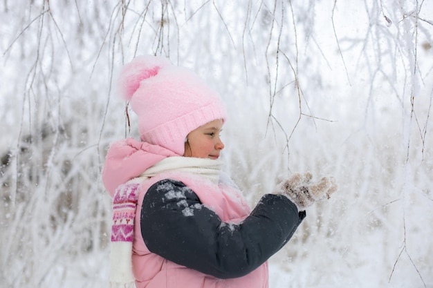 Kaukasisches junges Mädchen, das mit schneebedeckten Ästen spielt, die warme Winterkleidung tragen, die Hände betrachtet und mit Bäumen im Hintergrund lächelt Winterspaziergang im Wald