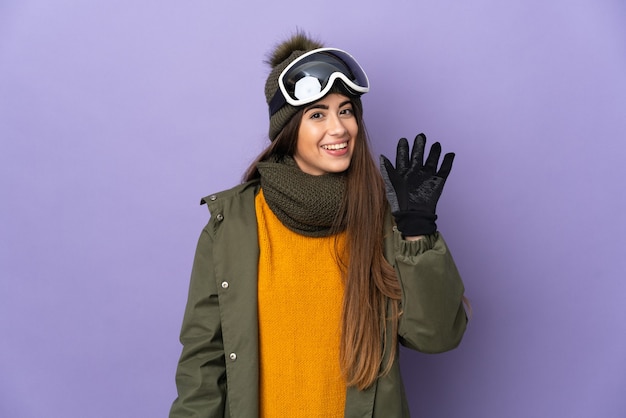 Kaukasisches Mädchen des Skifahrers mit Snowboardbrille lokalisiert auf lila Wand, die mit Hand mit glücklichem Ausdruck salutiert