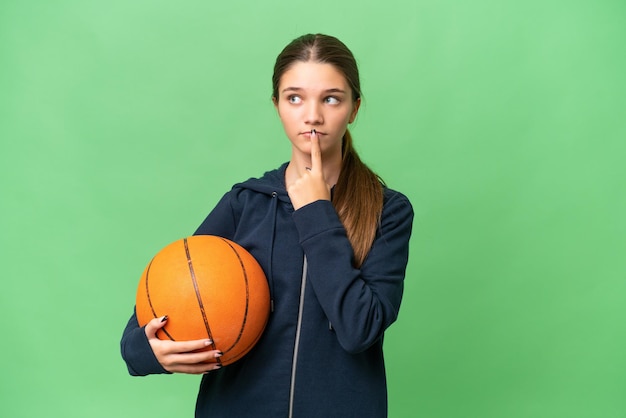 Kaukasisches Mädchen im Teenageralter, das Basketball vor isoliertem Hintergrund spielt und beim Aufblicken Zweifel hat