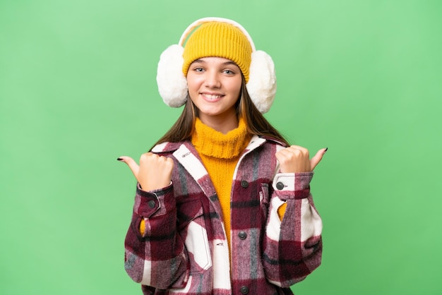 Kaukasisches Mädchen im Teenageralter, das Wintermuffs über isoliertem Hintergrund trägt, mit Daumen nach oben, Geste und Lächeln