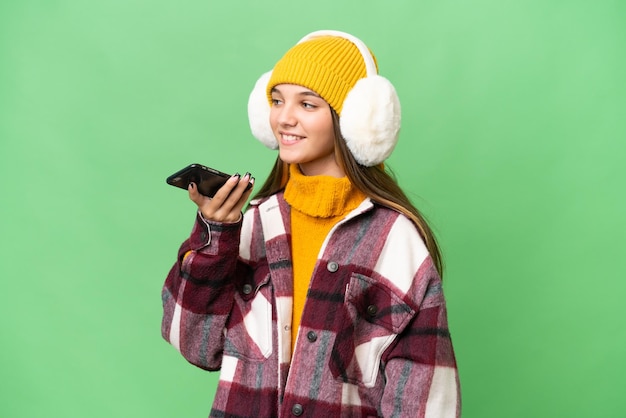 Kaukasisches Mädchen im Teenageralter, das Wintermuffs über isoliertem Hintergrund trägt und ein Gespräch mit dem Mobiltelefon führt