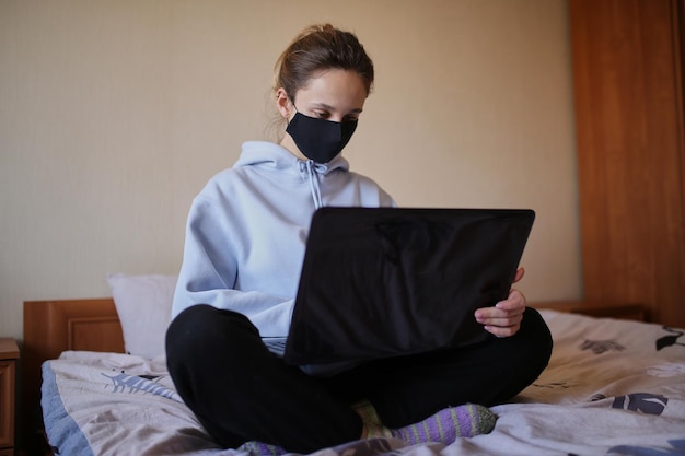 Kaukasisches Mädchen in blauem Hoodie und schwarzer Maske arbeitet isoliert am Laptop zu Hause